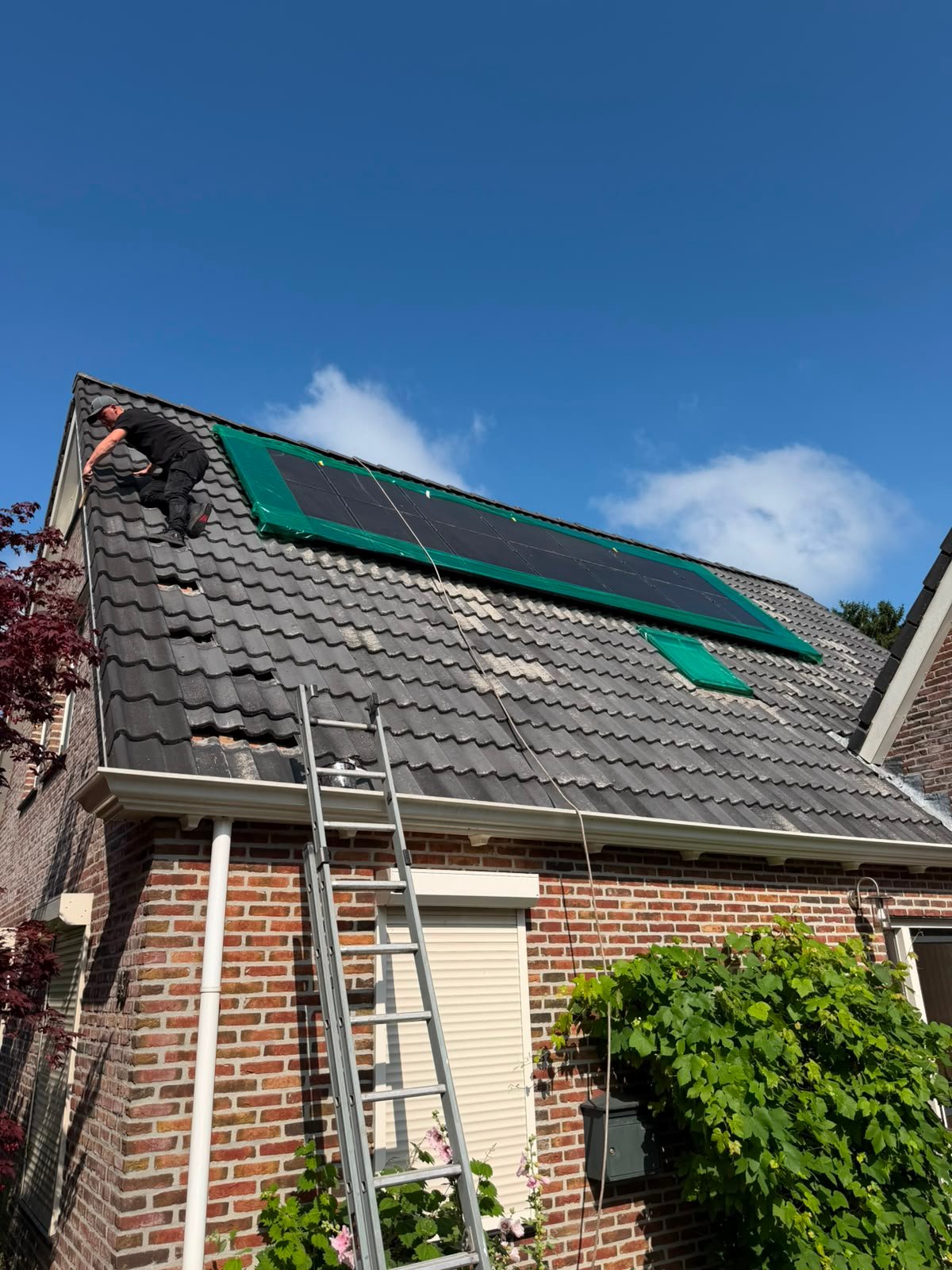 Betonpannendak tijdens het coaten: links ligt de coating er al op, rechts nog niet. Iemand werkt aan de dakrand en de zonnepanelen zijn afgeplakt met groene folie.