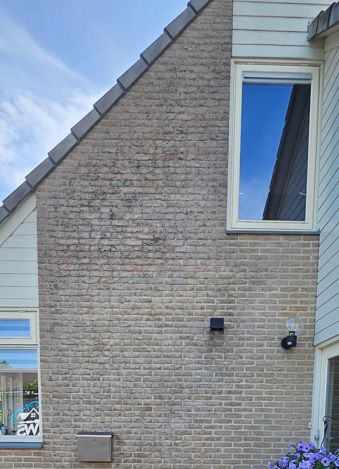 Voor: gele bakstenen puntgevel met raam en buitenlamp, grauw van grijze aanslag en een groene waas