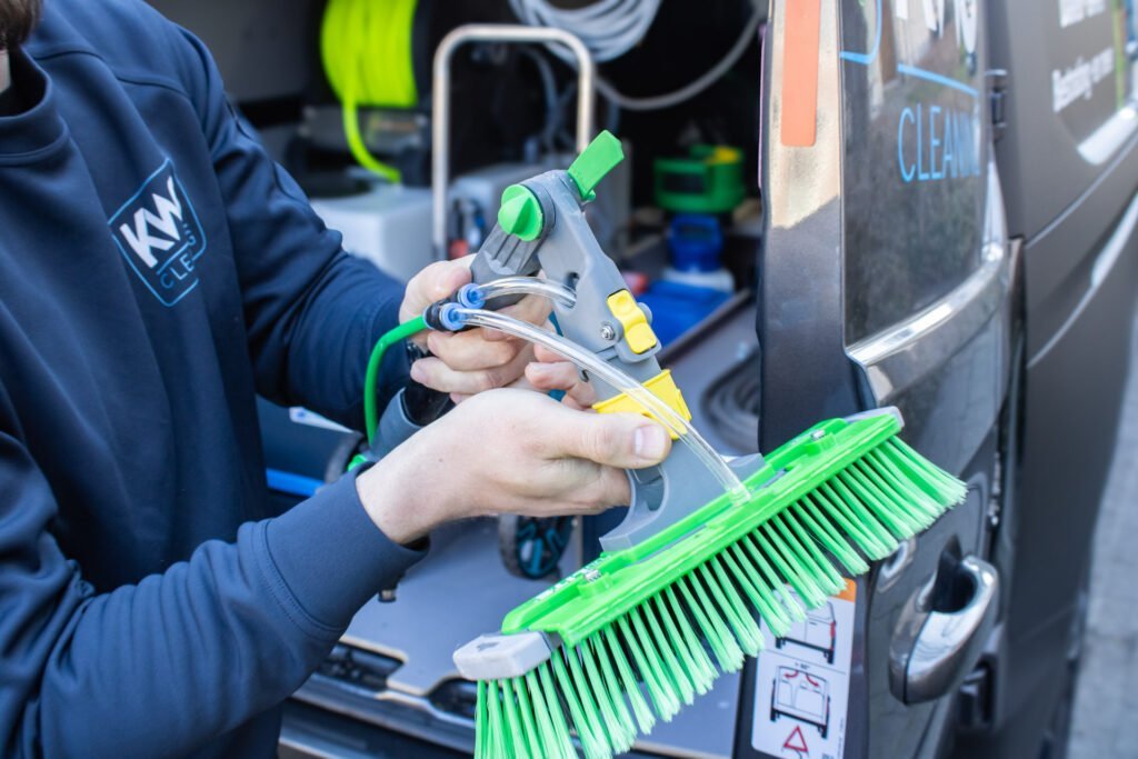 Handen met een borstel en een spuitpistool aan een slang, bij de bus met reinigingsmateriaal