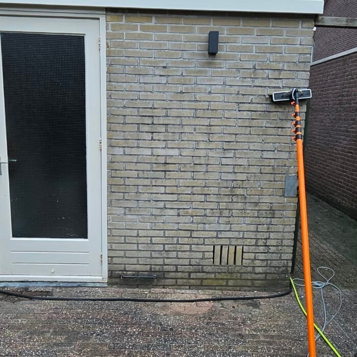 Borstel op een oranje telescoopsteel tegen een lichte bakstenen muur, met een tuinslang op het terras
